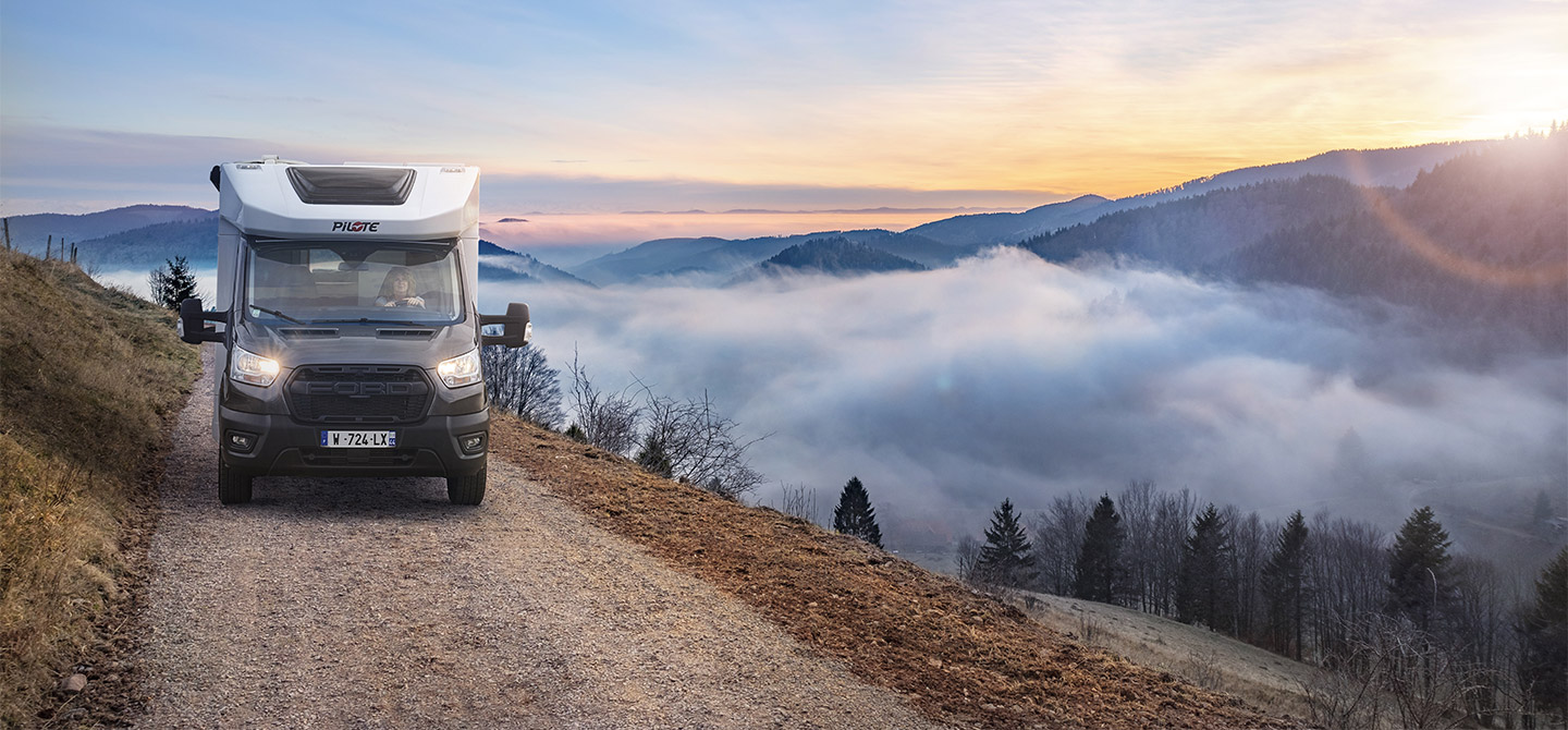 Camping-car profilé étroit Pilote roulant sur un chemin d'altitude, les nuages accrochés aux montagnes et le soleil levant.