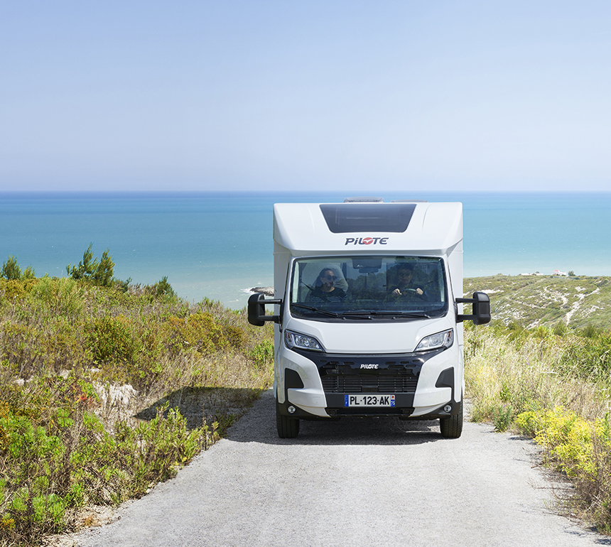 Camping-car profilé Pilote sur une route verdoyante de bord de mer, la mer turquoise au fond.