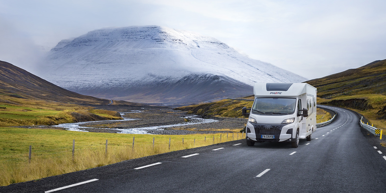 Camping-car profilé Pilote roulant dans un paysage d'Islande, entre végétations jaunies, cours d'eau et montagne enneigée.