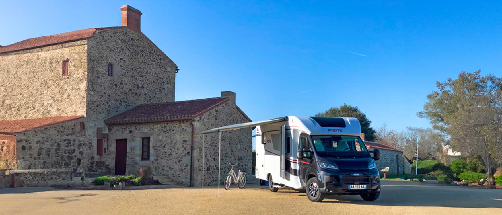 Camping-car profilé P720FC Sélection Pilote vu de côté, store banne déplié, à côté d'une bâtisse en pierres.